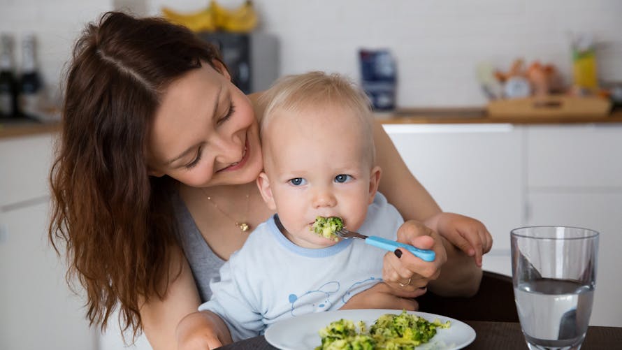 bébé mangeant des brocolis sur les genoux de sa maman