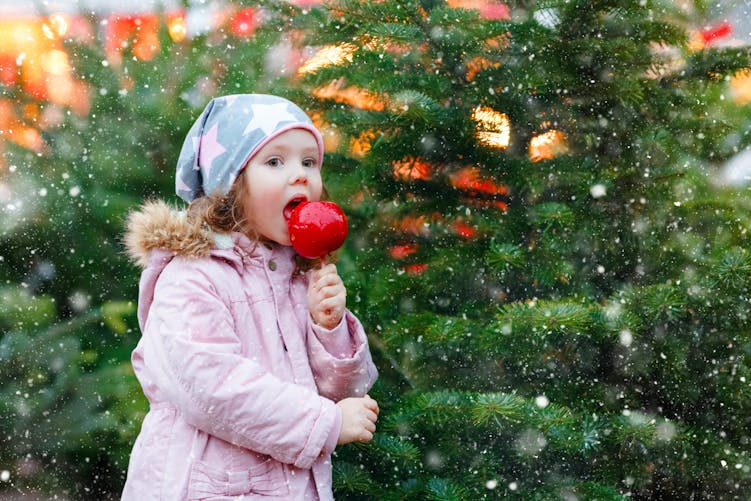 Une petite fille mange une pomme d'amour devant des sapins de Noël