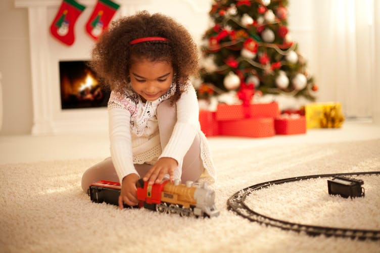 Une petite fille joue avec son train, devant un sapin de Noël.