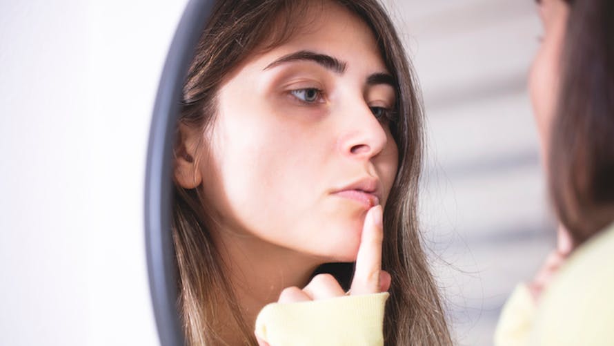 Une femme regarde un bouton de fièvre dans un miroir.