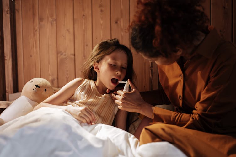Une petite fille est allongée dans un lit et une dame lui met du spray dans la bouche. 