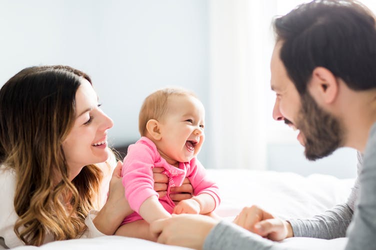 Deux adultes rigolent avec un bébé sur un lit.