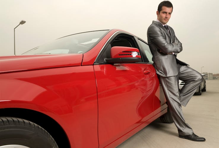 Un homme en costume gris s'adosse fièrement à sa voiture de luxe rouge