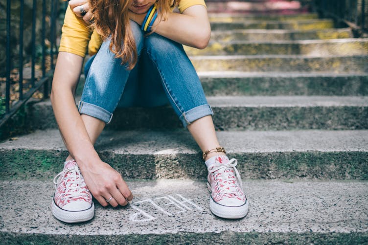 Une jeune fille, assise sur des marches, écrit "help" à la craie