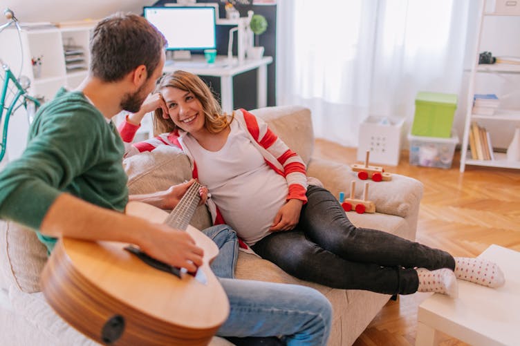 Sur un canapé, une femme enceinte écoute avec joie son ami jouer de la guitare