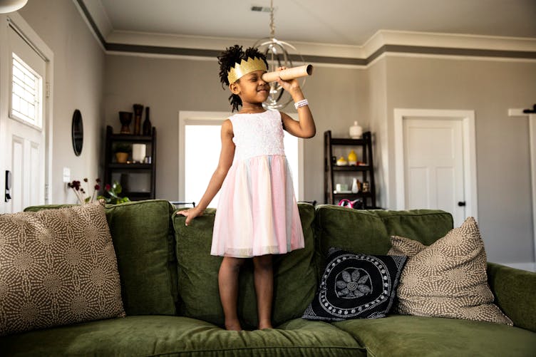 Une petite fille joue à la princesse en robe debout sur son canapé