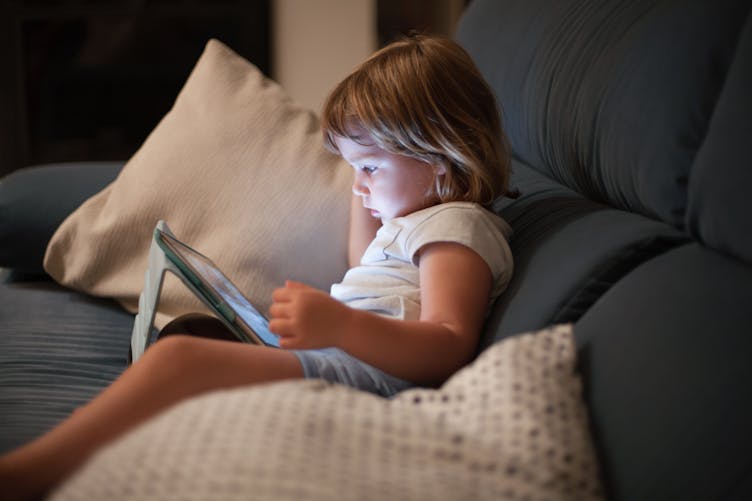Un enfant regarde l'écran d'un ipad en étant assis dans un canapé.