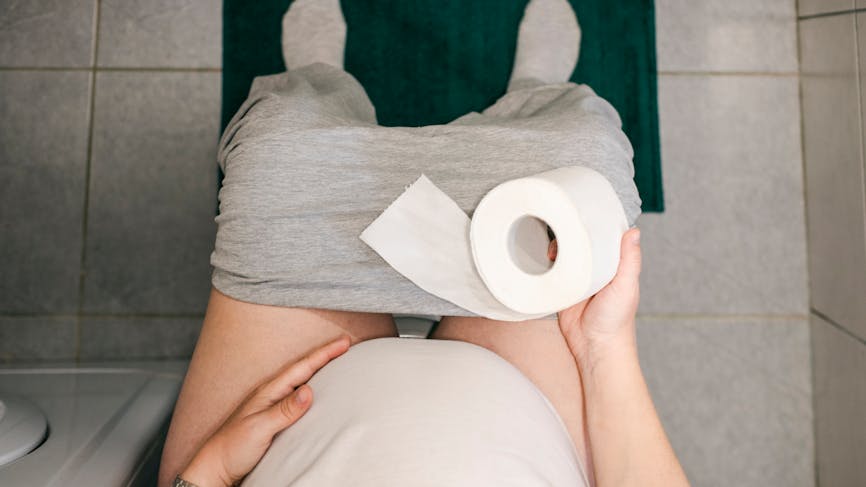 Une femme enceinte assise sur la cuvette des toilettes, tenant un rouleau de papier toilette