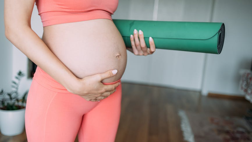 femme enceinte en tenue de sport tenant son ventre et un tapis de yoga