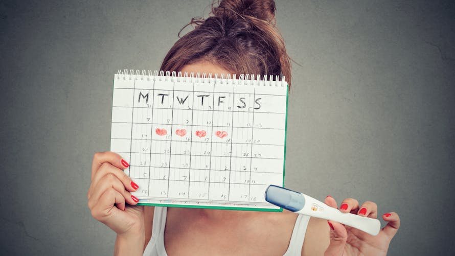 une femme se cachant derrière un calendrier pour suivre son cycle menstruel, tenant un test de grossesse positif à la main.
