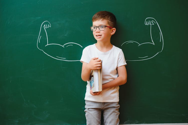 Un jeune garçon à lunettes pose, gêné, devant un tableau noir où des bras musclés sont dessinés à la craie