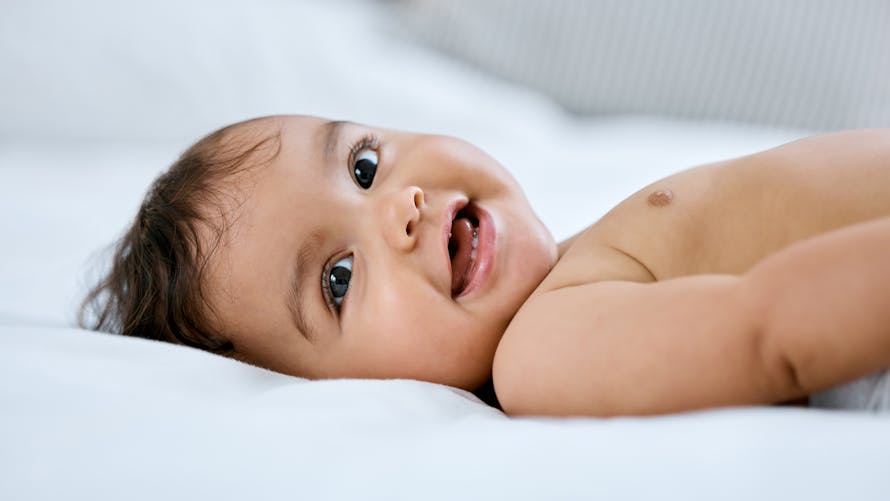 Un bébé aux cheveux bruns sourit sur un lit. 