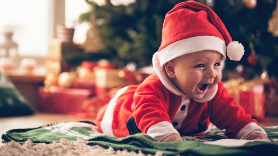 Bébé déguisé en Père Noël allongé sur un tapis avec des cadeaux en arrière-plan.